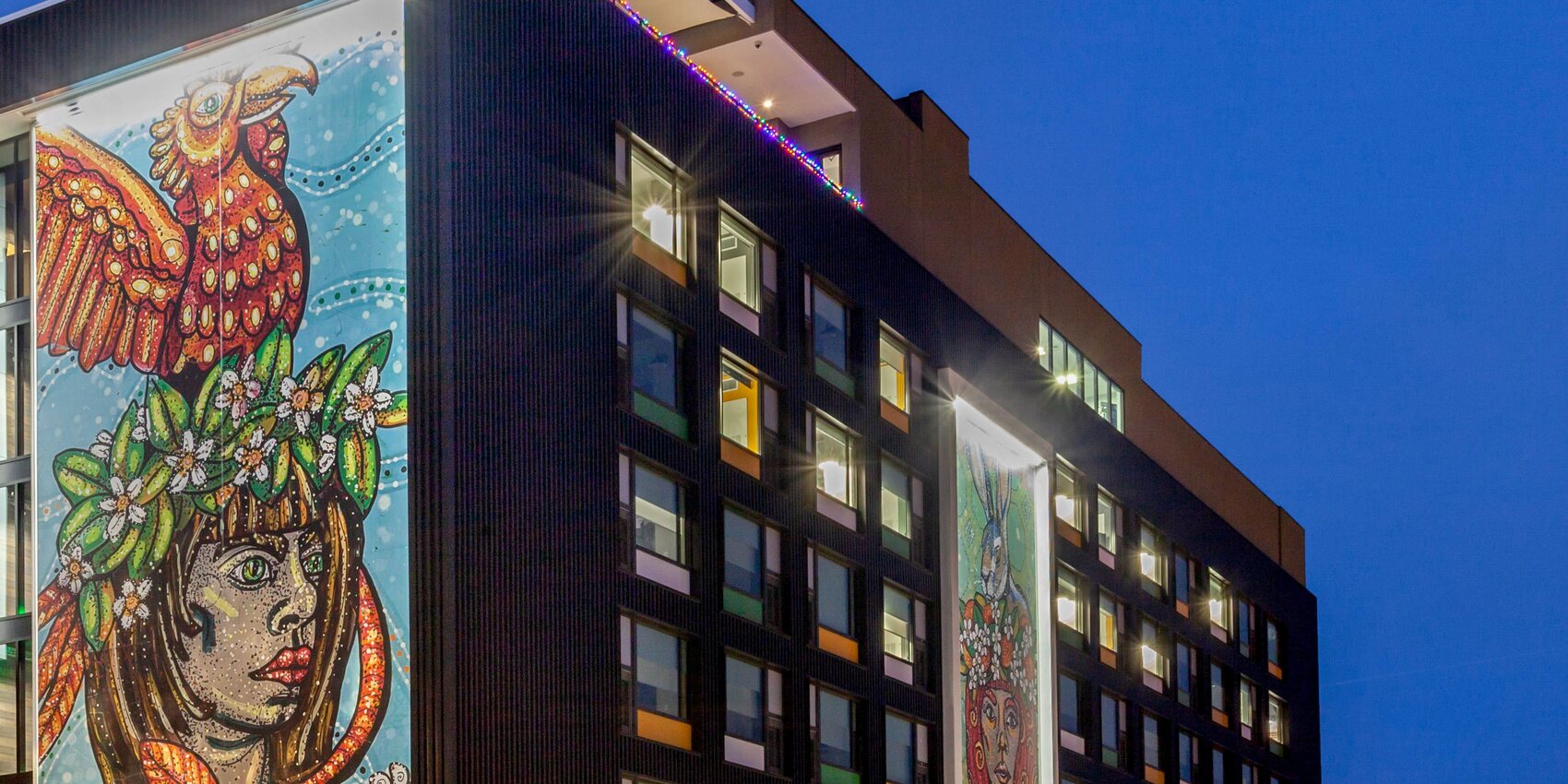 The hotel at dusk, with a six storey mural of a woman crowned in orange blossom